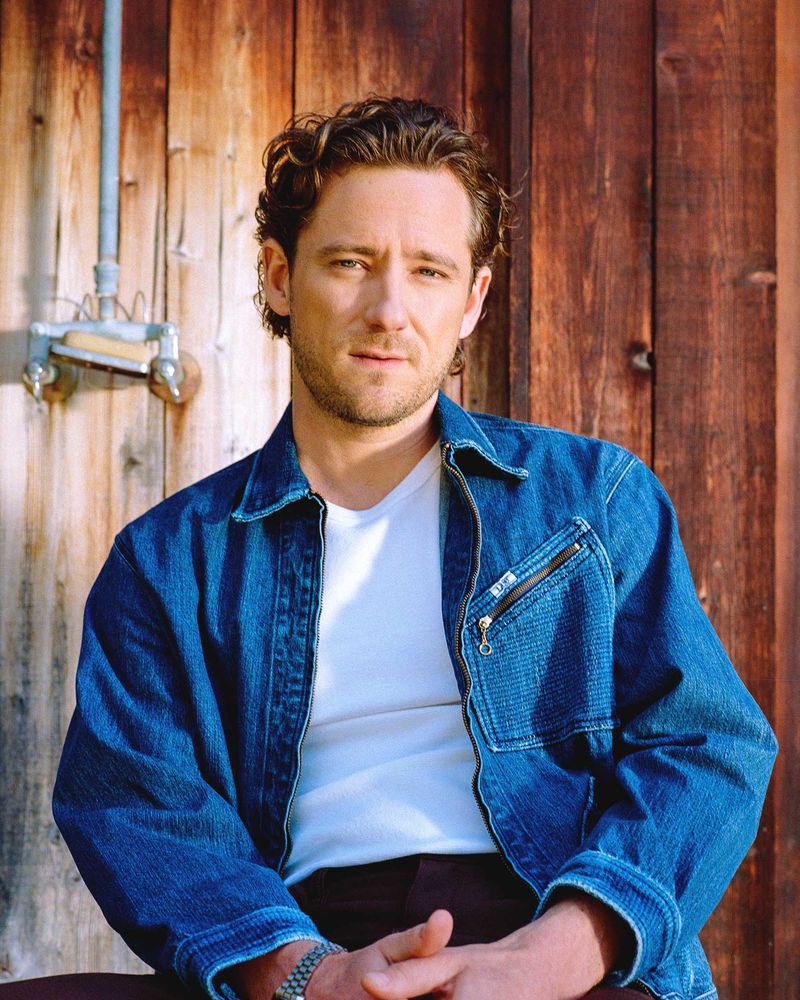 Portrait of an adult male with curly brown hair wearing a blue denim jacket and white t-shirt posing in front of a wooden wall.