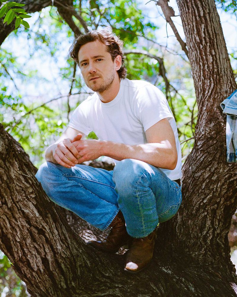 Portrait of an adult male crouching on a tree branch wearing jeans, a white t-shirt and cowboy boots outdoors during daytime.