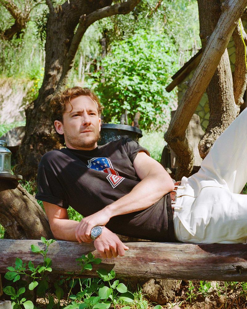 Adult male laying on a wooden log in a garden wearing a graphic tee and white pants, with green trees and a small house in the background.