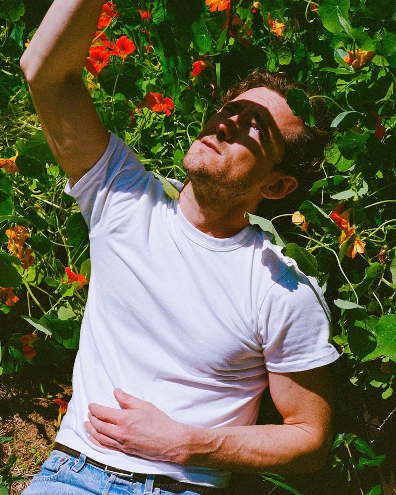 Young adult man lies in a field of orange flowers and green foliage, wearing a white tee and blue jeans, looking up into sunlight.