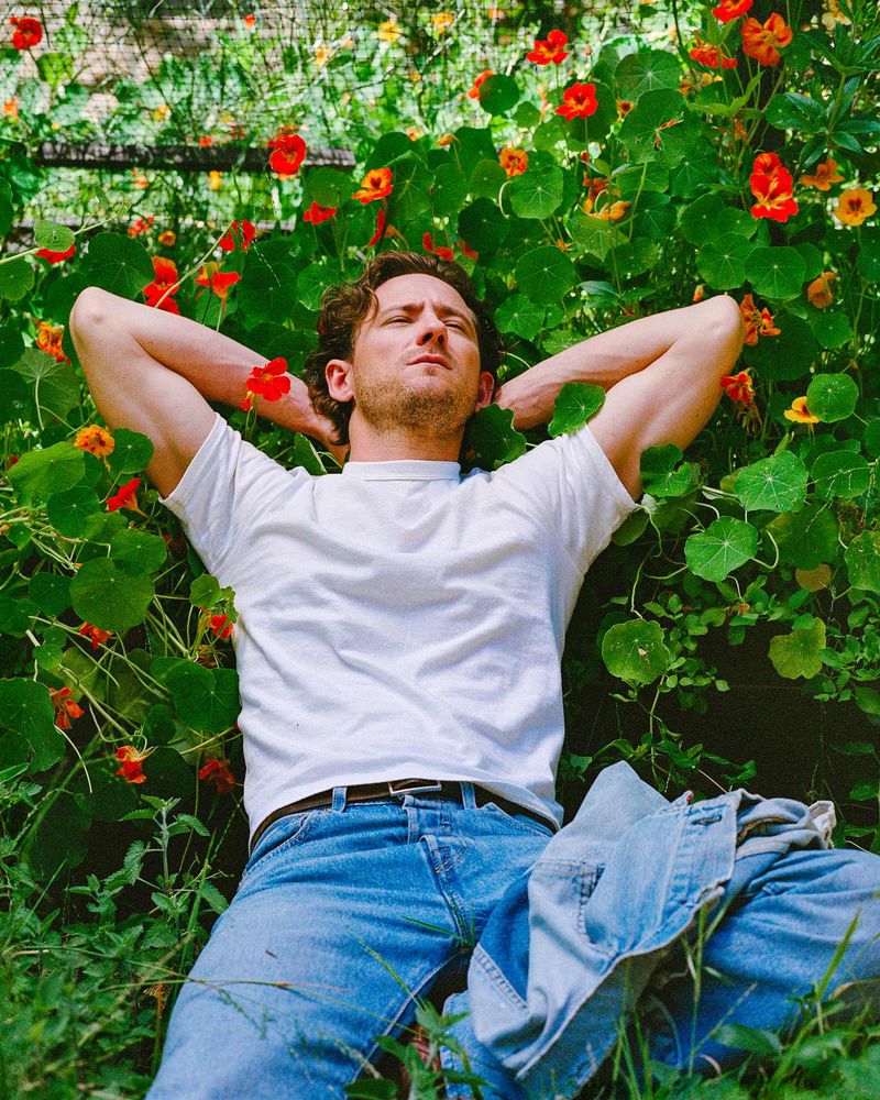 Man in a white T-shirt and jeans lying in a field of flowers with his arms behind his head, looking relaxed and peaceful in natural light.