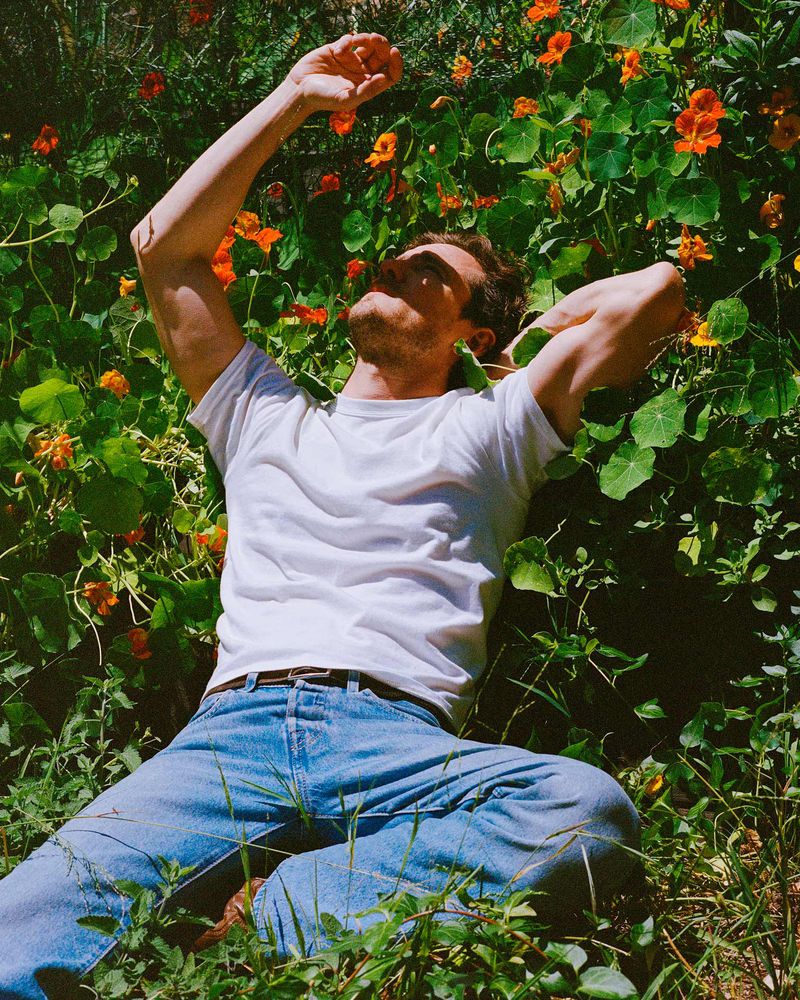 Young adult male in jeans and a white t-shirt lying on his back in bright sunlight, surrounded by lush green foliage and orange wildflowers.