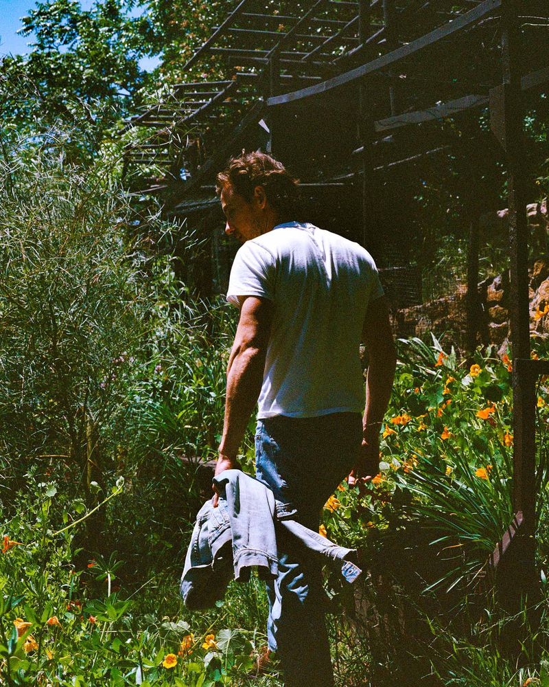 Adult male in jeans and white t-shirt stands in a garden with flowers, holding a denim jacket, with foliage and a metal pergola.