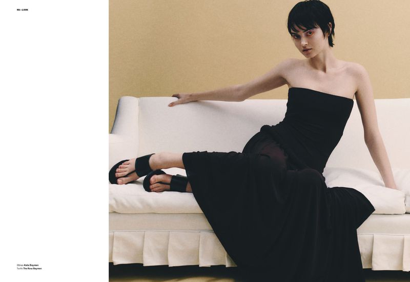 Fashion editorial shot of a woman in a black dress lounging on a white sofa against a tan backdrop in a minimalist, indoor setting.
