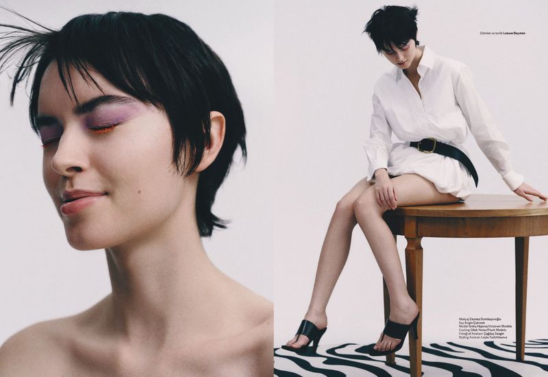A woman with short dark hair and purple eyeshadow sits on a wooden table in front of a white background. She wears a white shirt.