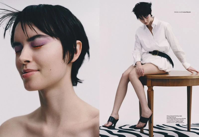 A woman with short dark hair and purple eyeshadow sits on a wooden table in front of a white background. She wears a white shirt.