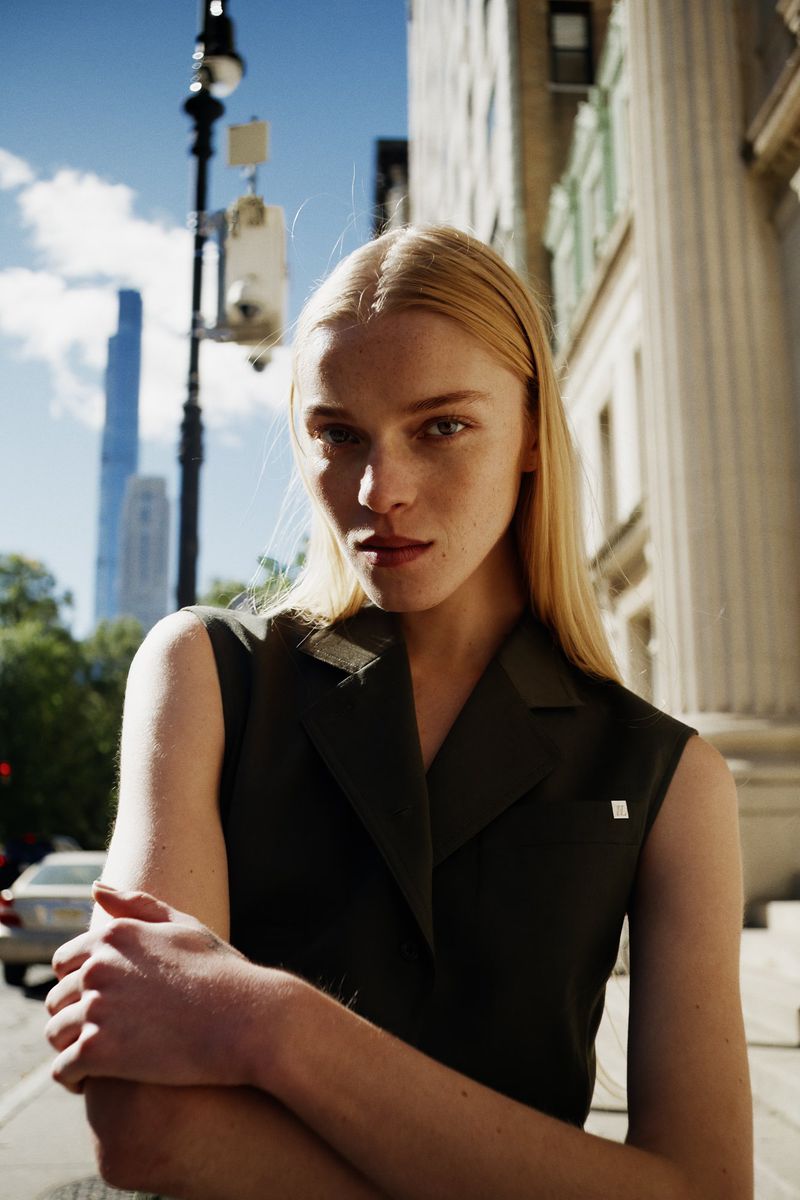 Portrait of a young woman with blonde hair wearing a dark sleeveless top standing in an urban environment with tall buildings in the background.