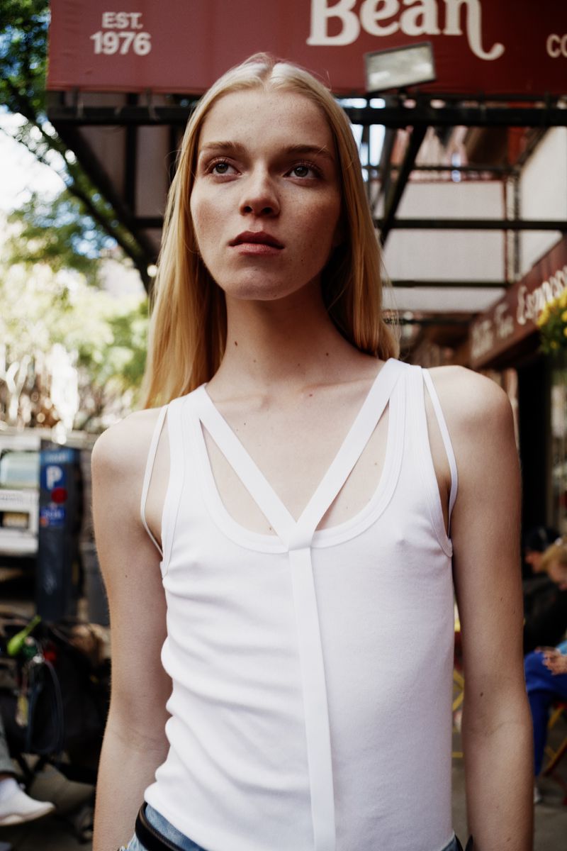 A woman with blonde hair is standing outside a cafe. She's wearing a white tank top with a V-shaped strap design and blue jeans.