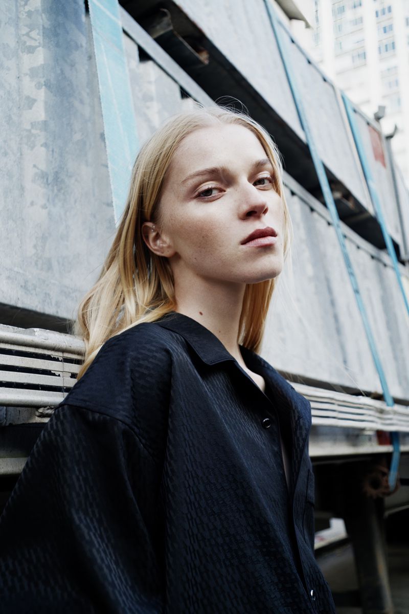 Portrait of a blonde adult woman wearing a black collared shirt standing outside near a textured metal wall in daytime, medium shot.