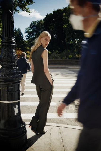Blonde woman in a dark green suit glances over her shoulder while walking across a crosswalk in the city on a sunny day.