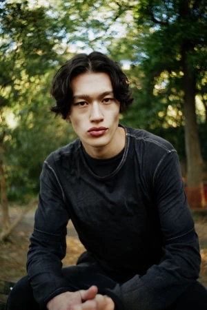 Portrait of a male model wearing a dark long-sleeve top outdoors against a backdrop of trees in a park.