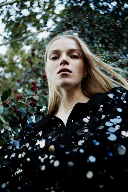 Eye-level shot of a blonde woman looking directly at the camera, wearing a dark sequined top, with a leafy green background.
