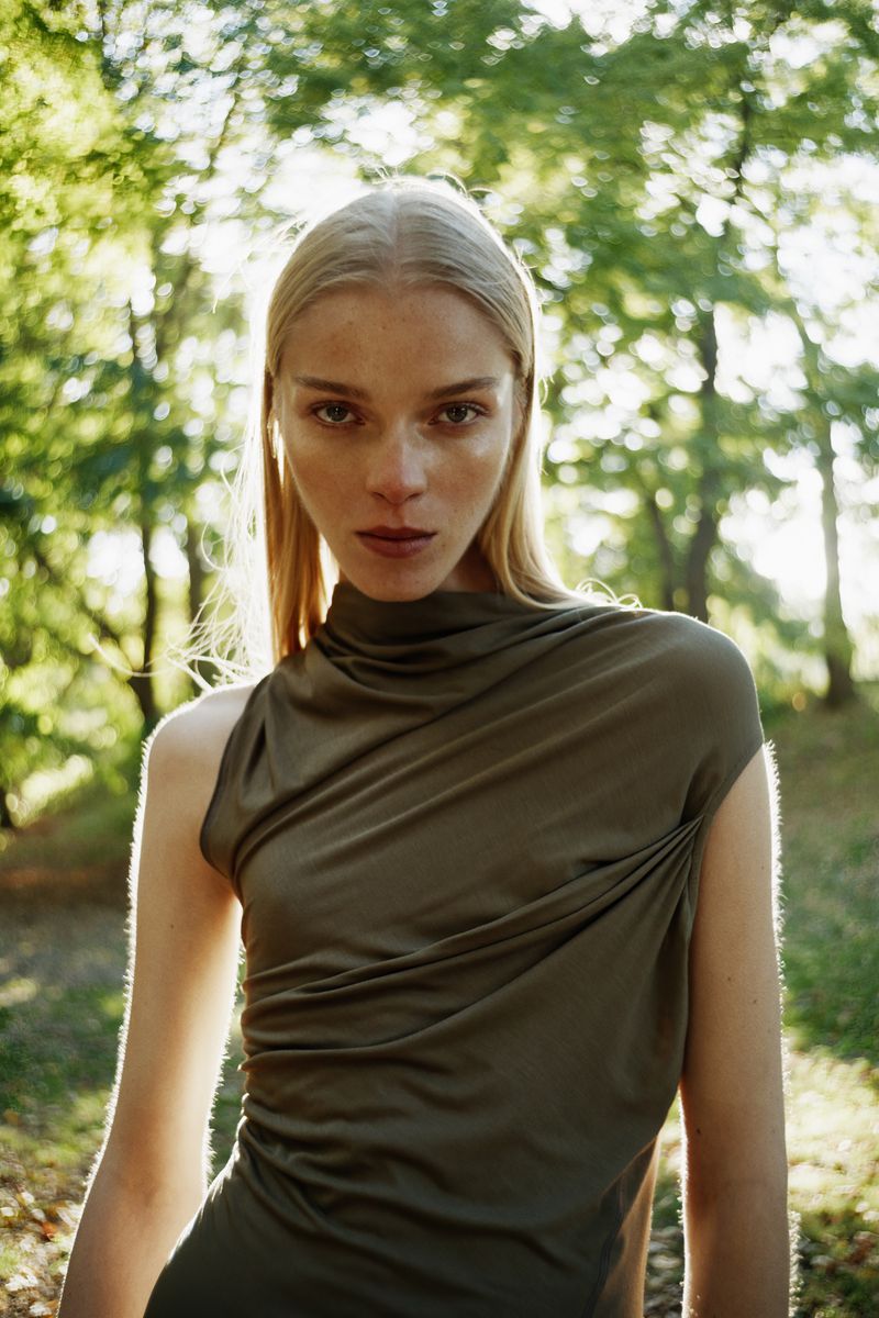 Model with long blonde hair wearing an olive green sleeveless top outdoors against a backdrop of trees