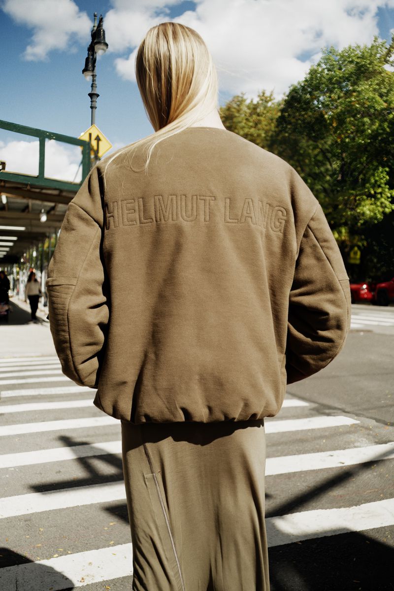 Model seen from behind wearing an olive green crewneck sweatshirt with embossed Helmut Lang branding crossing a city crosswalk.