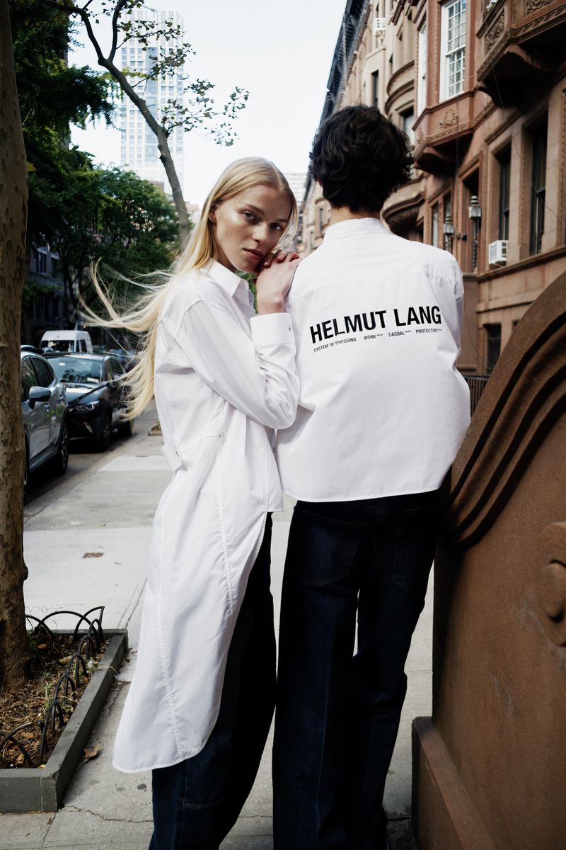 Two fair-skinned adult models in white button-down shirts and dark jeans stand on a city sidewalk with residential buildings in the background.
