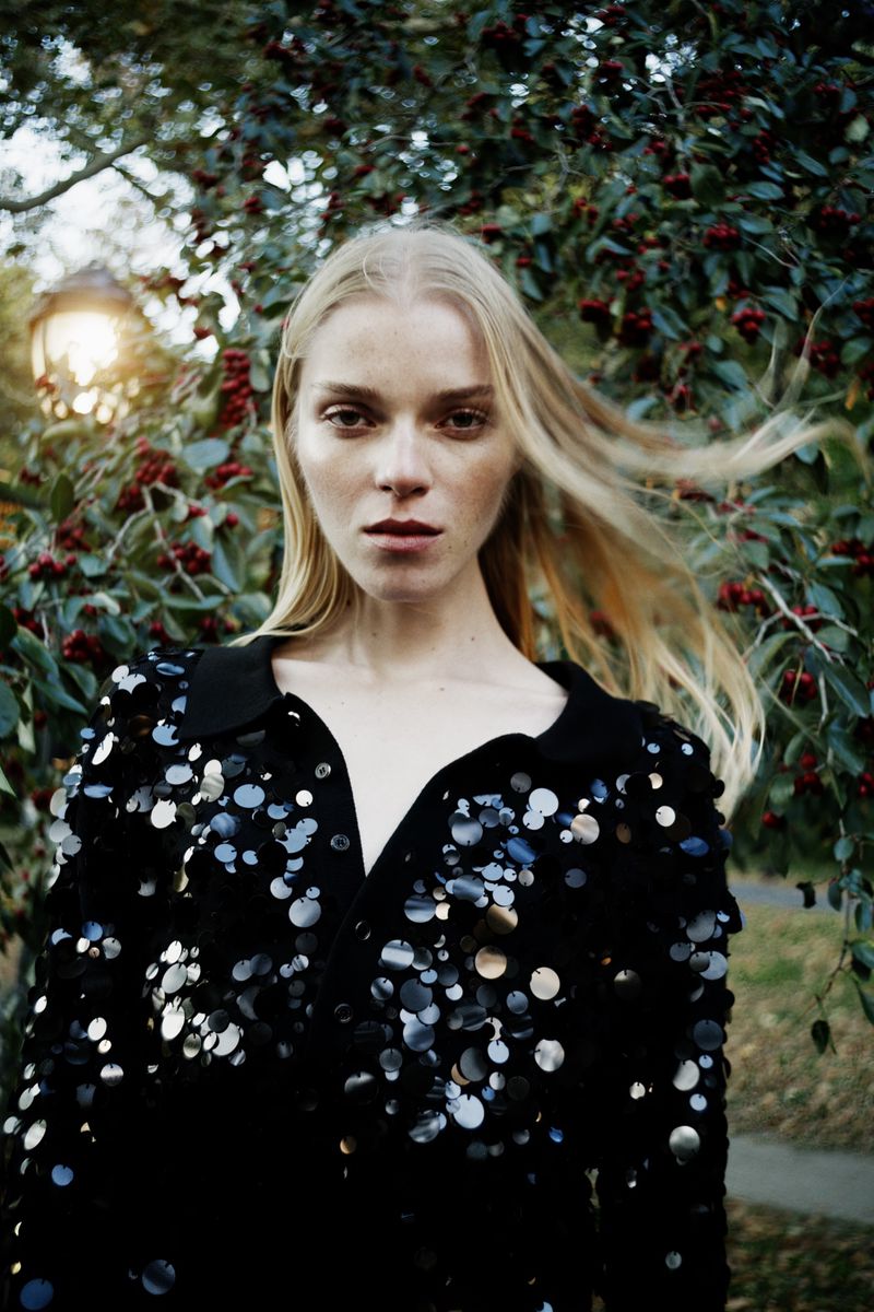Model with long blonde hair wearing a black sequined top outdoors against a leafy background with red berries.