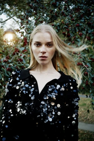 Model with long blonde hair wearing a black sequined top outdoors against a leafy background with red berries.