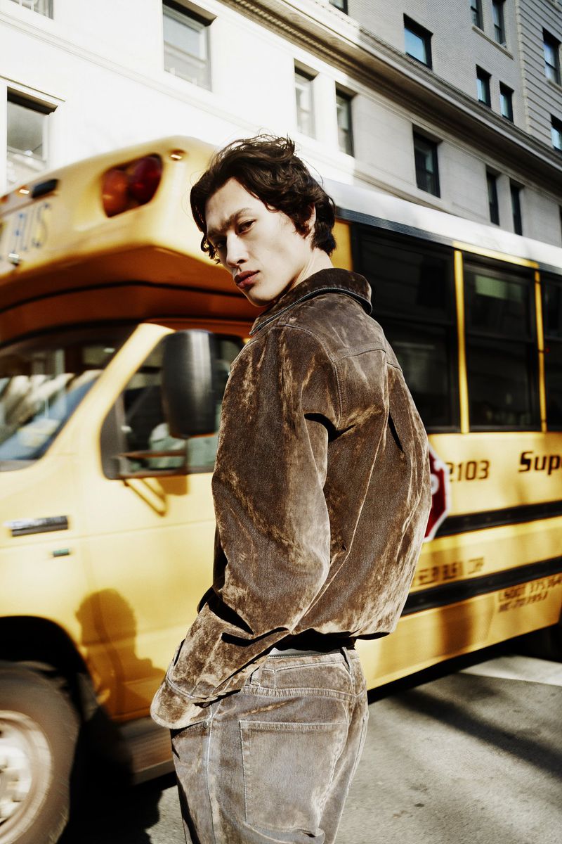 Young adult man in brown jacket poses in front of a yellow school bus with a confident expression in an urban setting outdoors.