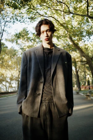 Model wearing a grey blazer and dark trousers standing outdoors on a paved path lined with trees.