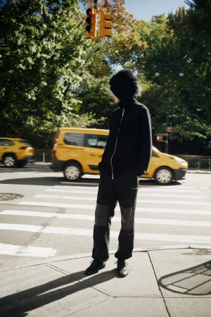 Model wearing a black hooded jacket and pants standing on a city street corner with yellow taxis passing in the background.