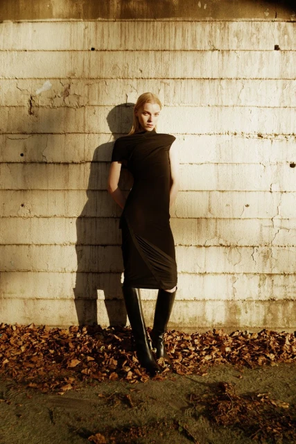 A blonde female fashion model in a black dress poses against a textured wall, knee-high boots, fallen leaves at her feet, outdoor fashion editorial.