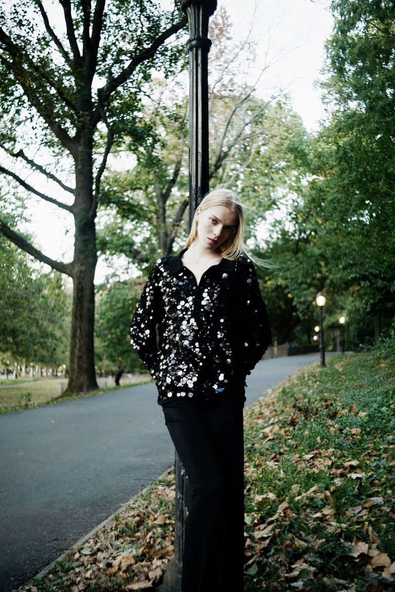 Woman in a sequined jacket poses in a park with fall foliage. A lamppost is in the scene as the woman stands on a walking path.