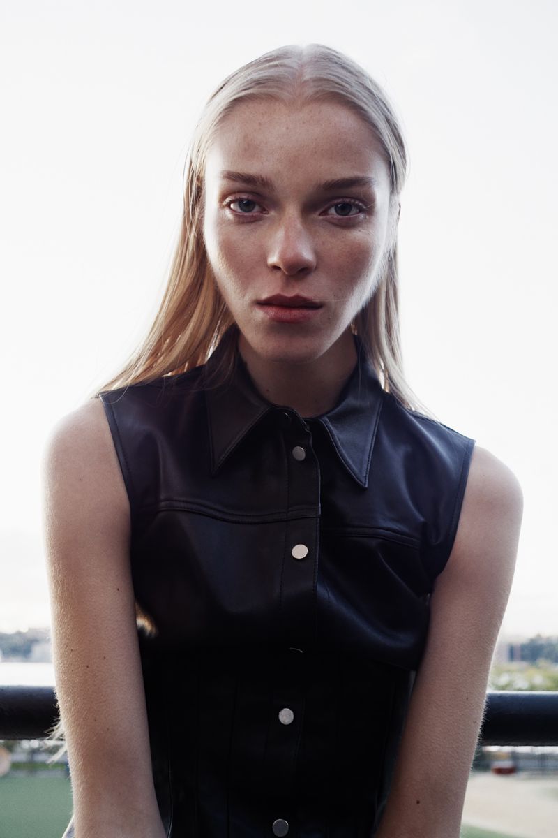 Portrait of a young woman with long blonde hair wearing a dark, sleeveless top with silver buttons, gazing intently at the camera outdoors.
