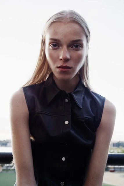 Portrait of a young woman with long blonde hair wearing a dark, sleeveless top with silver buttons, gazing intently at the camera outdoors.