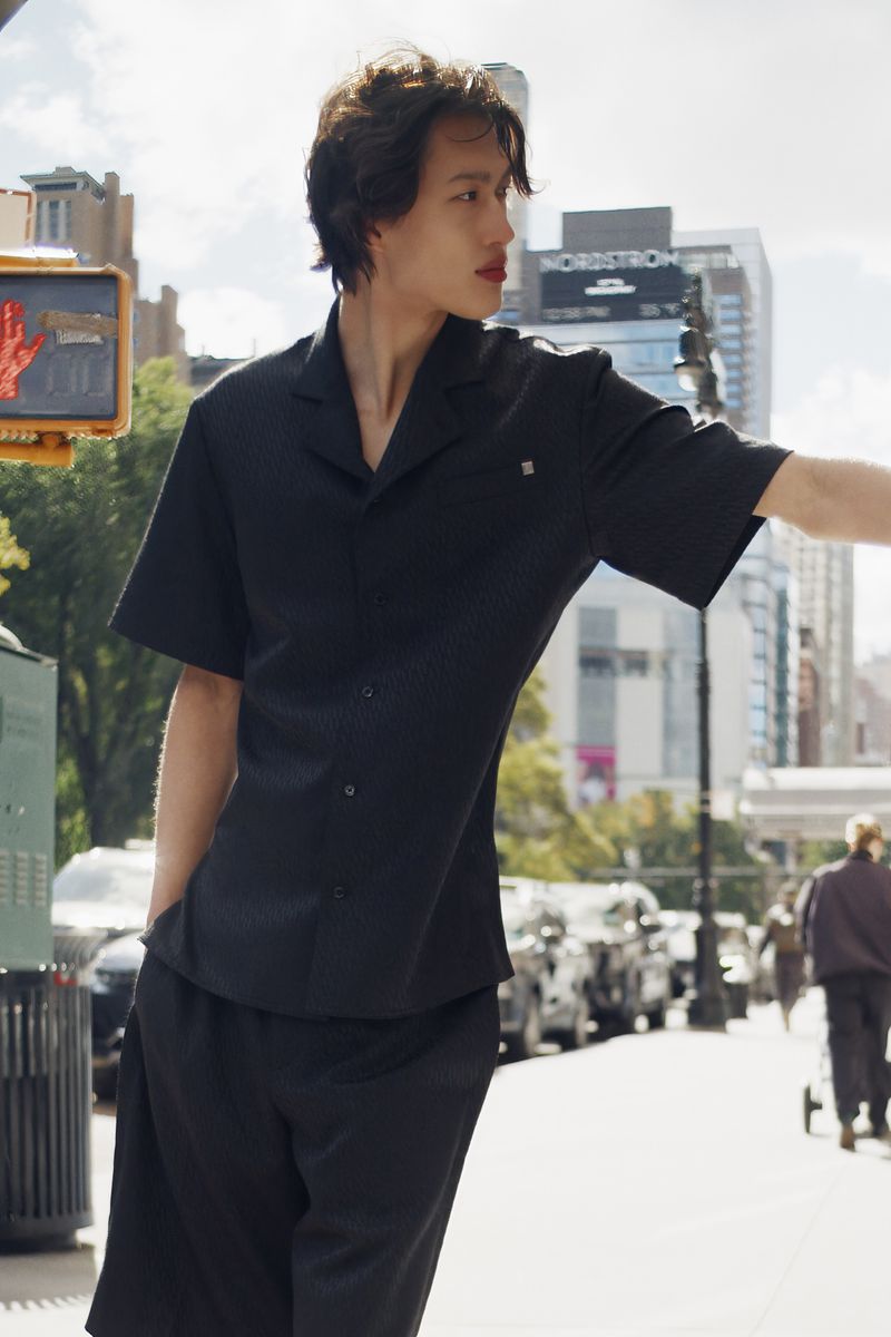 Adult male in black short-sleeve button-down shirt and matching shorts stands on sidewalk with city buildings in the background.