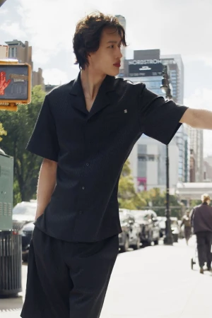 Adult male in black short-sleeve button-down shirt and matching shorts stands on sidewalk with city buildings in the background.