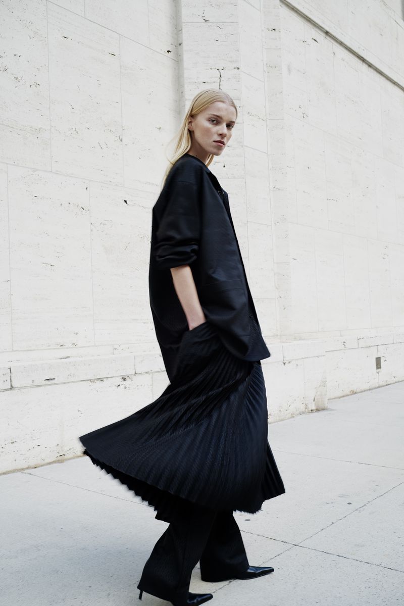 Fashionable woman in a black jacket and pleated pants standing against a textured white wall in natural outdoor lighting.