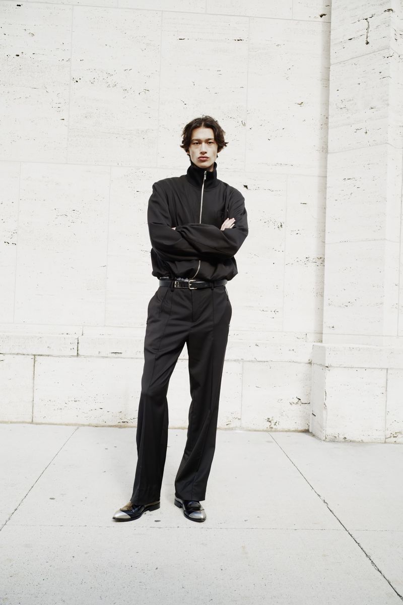 Full-length portrait of a young adult male with crossed arms, wearing a black jacket and dress pants against a textured wall.