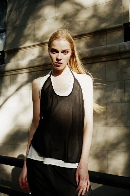 Outdoor shot of a blonde woman in a black top, standing against a light stone wall with dappled sunlight, looking at the camera.