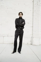 Male model in a black zip-up jacket and trousers standing with arms crossed against a pale stone wall
