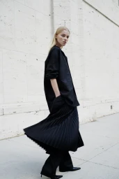 Model in a black shirt and pleated skirt standing against a light stone building exterior for Helmut Lang