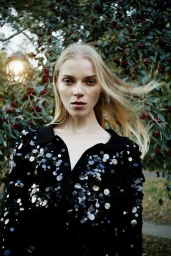 Model with long blonde hair wearing a black sequined top outdoors against a leafy background with red berries.