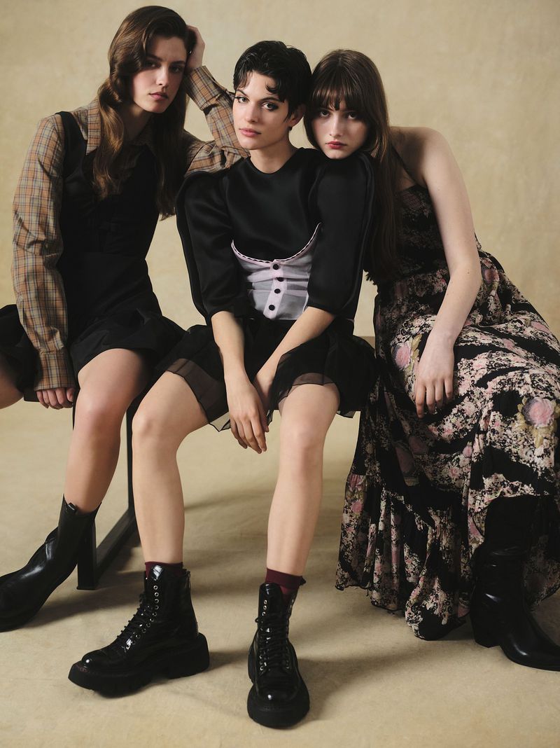 Three young women pose indoors in a fashion shot, showcasing vintage-style dresses and glossy black boots against a neutral backdrop.