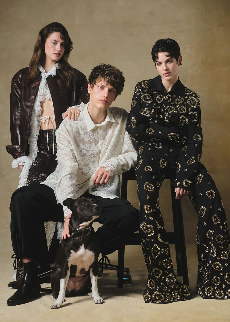 Three fashion models and a black and white dog pose indoors wearing patterned clothing in a studio setting with neutral backdrop.