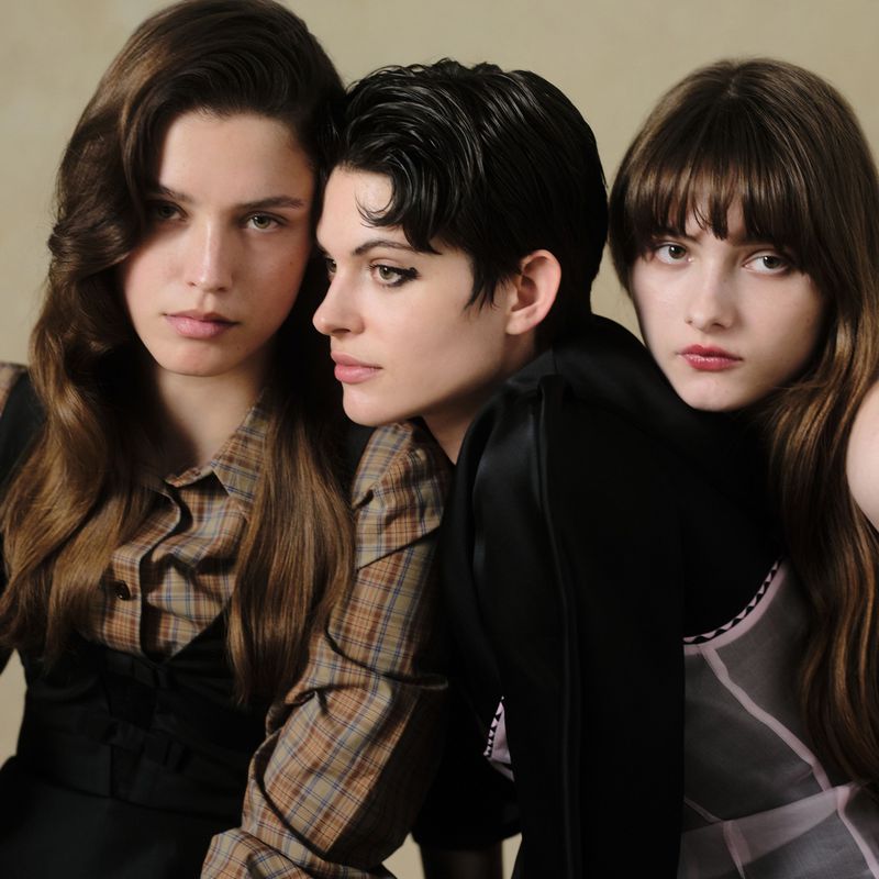 Three young women pose closely together indoors wearing fashionable tops. The image is lit with soft light, and has a natural look.