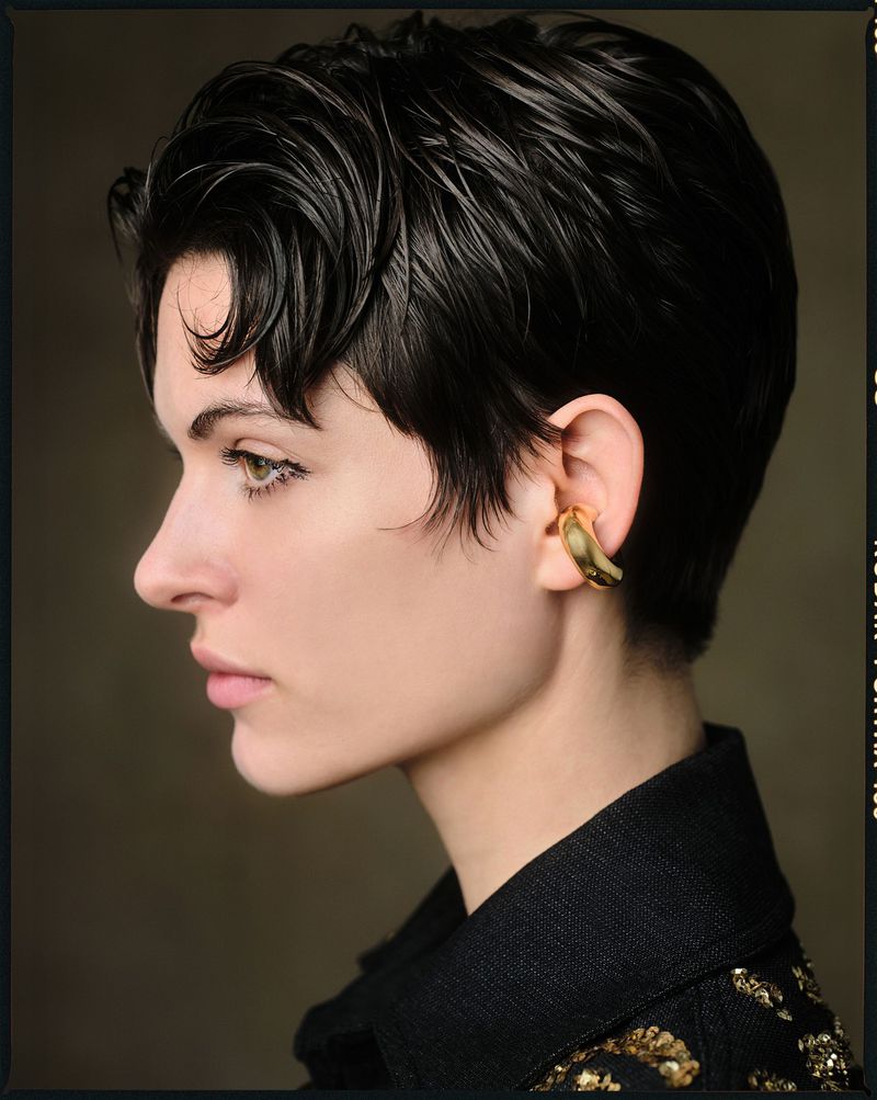 Portrait of a woman with dark hair and gold ear cuff. Close-up studio shot featuring makeup and elegant, minimalist styling.
