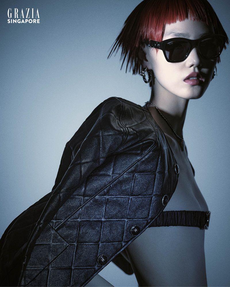 Fashion editorial shot of a red-haired adult woman in sunglasses and a black jacket posing against a cool-toned studio backdrop.