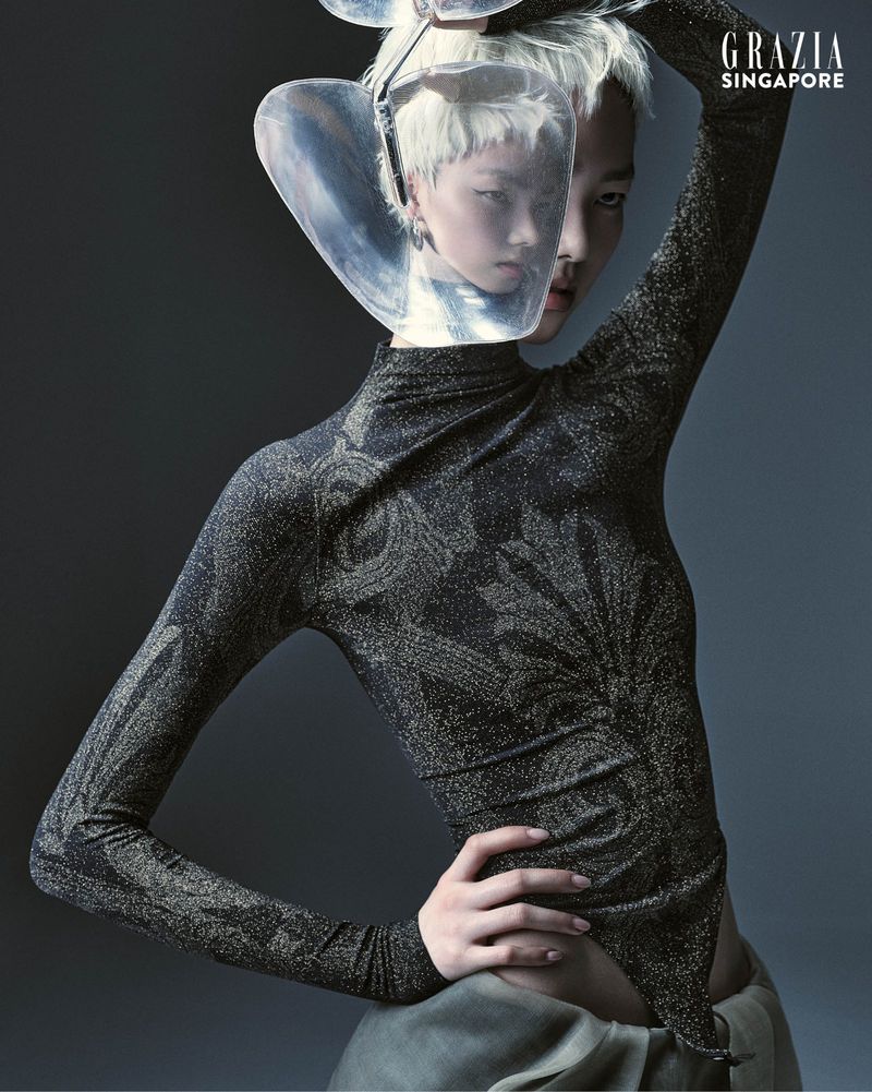 Fashion editorial shot of a woman with short white hair wearing a sparkling, patterned long-sleeve bodysuit and a clear visor headpiece.