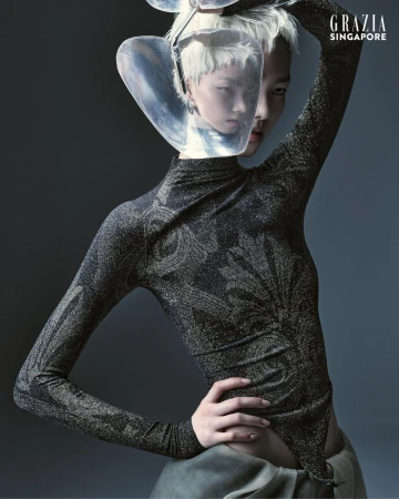 Fashion editorial shot of a woman with short white hair wearing a sparkling, patterned long-sleeve bodysuit and a clear visor headpiece.