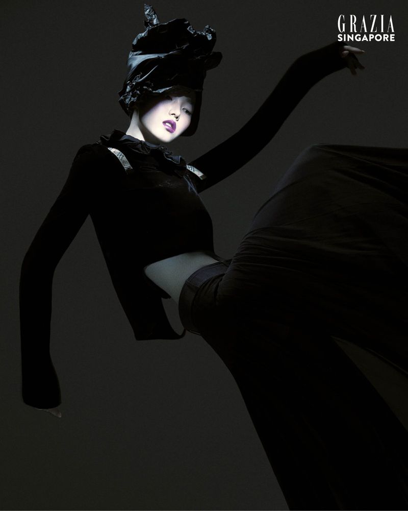 Fashion editorial shot of a woman wearing an all-black outfit and a hat, posing with arms upraised in a photo studio with black backdrop.