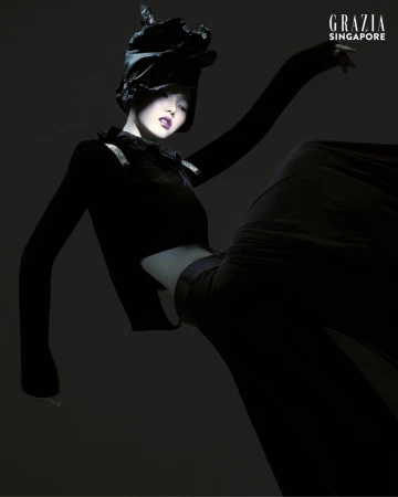 Fashion editorial shot of a woman wearing an all-black outfit and a hat, posing with arms upraised in a photo studio with black backdrop.
