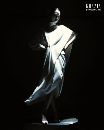 Model Jiahui Zhang wearing a sculptural draped white dress in dramatic spotlighting against a black background.