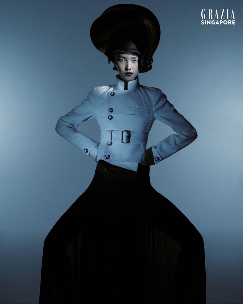Fashion editorial shot of an Asian model wearing a large black hat, light jacket, and long black skirt against a blue background.