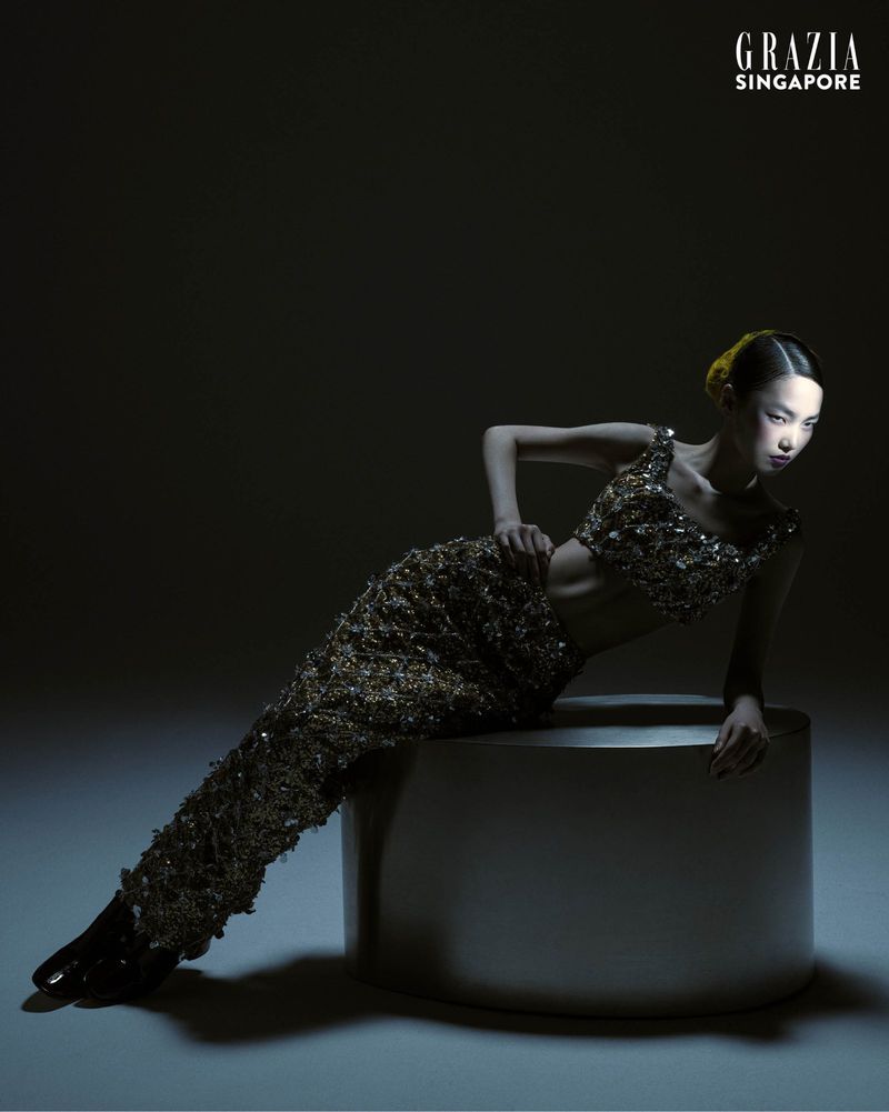 Glamour shot of an Asian female model in gold, sequined two-piece outfit, reclining on a round metal stool, against a dark background.