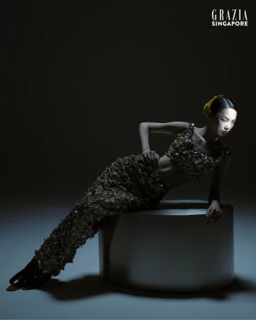 Glamour shot of an Asian female model in gold, sequined two-piece outfit, reclining on a round metal stool, against a dark background.
