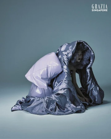 Fashion shot of a woman kneeling wearing a large, hooded, iridescent blue garment and a lavender pillow, against a gradient background.
